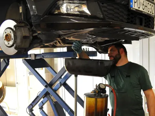 Mechanik w warsztacie wymienia zużyty olej samochodowy. Podniesiony pojazd i naczynie na płyn wskazują na rutynową czynność serwisową.