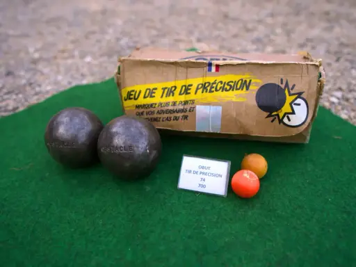 Deux boules de pétanque, un cochonnet et une boîte "Jeu de tir de précision" suggèrent un défi de poids et de diamètre pour le tireur.