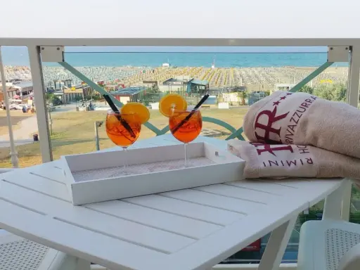 Due aperitivi arancioni e asciugamani con logo "Hotel Rivazzurra Rimini" sul tavolo, con vista sul rivazzurra mare e spiaggia attrezzata.