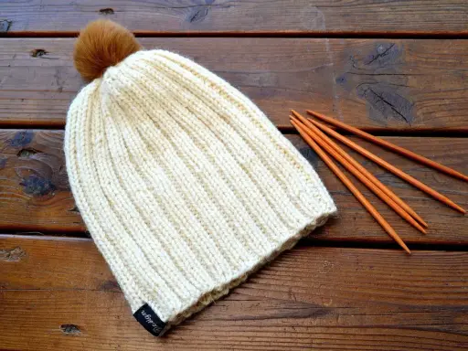 En ribbstickad mössa med en brun pompom ligger på ett träbord bredvid stickor. Perfekt för ett ribbstickad mössa gratis mönster.