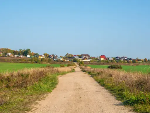 Droga gruntowa prowadzi do osiedla domów. Minimalna szerokość drogi dojazdowej do działki budowlanej jest tu widoczna, otoczona polami i trawami.