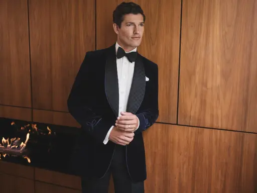 A man in a dark velvet tuxedo with a bow tie, embodying festive chic. He stands by a modern fireplace with flames.
