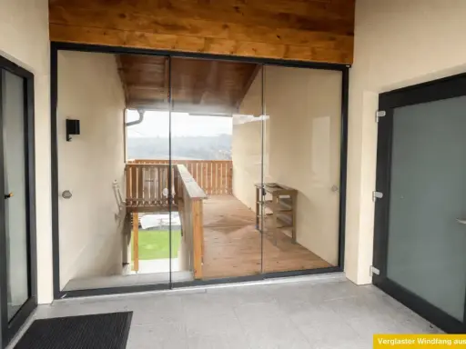 Moderner, verglaster Windfang aus Aluminium mit Holzbalken und Blick auf eine Holzterrasse.
