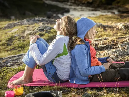 Två personer, en vuxen och ett barn, sitter rygg mot rygg på en liggunderlag i naturen. En härlig stund att vandra med barn.