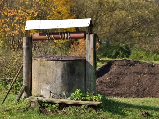 Stary, betonowy krąg studni kręgowej, z drewnianą konstrukcją i metalową korbą, otoczony jesienną zielenią.