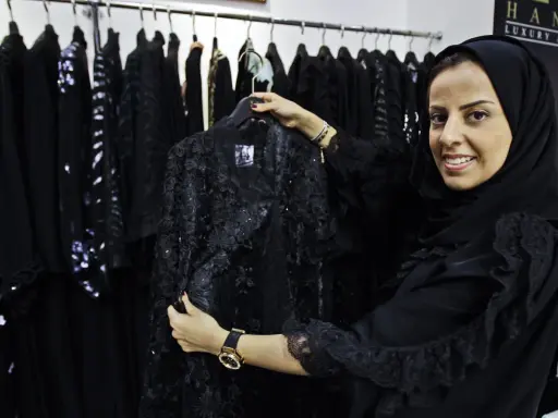 A woman smiles, holding a black lace abaya, showcasing elegant Saudi Arabia fashion.