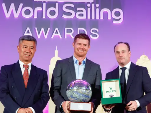 Tom Slingsby mit Auszeichnung bei den World Sailing Awards. Er hält eine Trophäe, daneben zwei Männer mit Geschenken.
