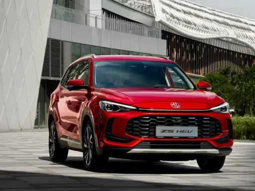 Czerwony MG ZS HEV, nowoczesny SUV hybrydowy, zaparkowany przed futurystycznym budynkiem.