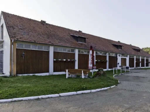 Długi budynek stajni z drewnianymi drzwiami i oknami, z parasolem i ławką przed nim.