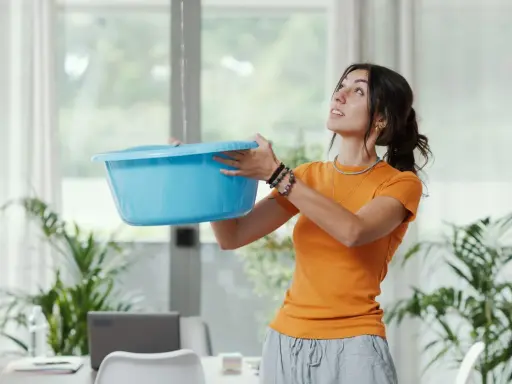 Une femme tente de contenir une fuite d'eau avec un bac bleu, visiblement exaspérée par le dégât des eaux voisin récalcitrant.