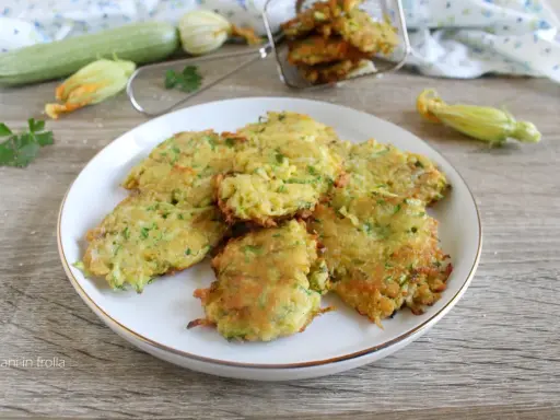 Buñuelos de calabacín dorados y crujientes en un plato blanco, con calabacines frescos y flores en el fondo.