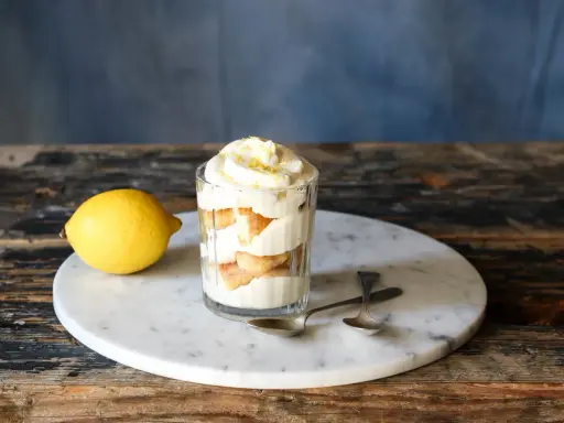 Ein Glas mit geschichtetem Zitronen Tiramisu, garniert mit Zitronenschale. Daneben liegt eine Zitrone.