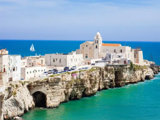 Un borgo bianco su scogliere a picco sul mare turchese, con una chiesa imponente. Un assaggio di cosa vedere in Puglia, con un veliero all'orizzonte.