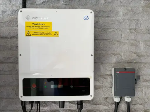 En vit GoodWe växelriktare 10kW monterad på en grå stenvägg. Bredvid sitter en grå ABB-box.