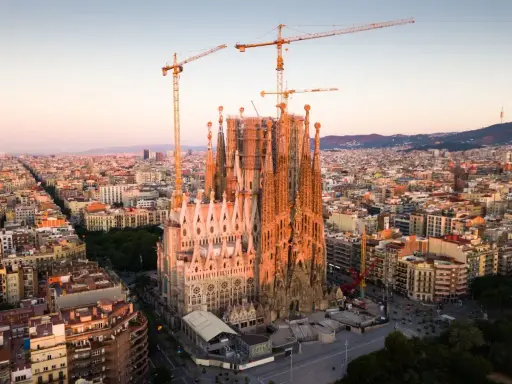 Widok z lotu ptaka na Sagrada Familia w Barcelonie, wciąż w budowie, z dźwigami górującymi nad miastem. Słońce oświetla fasadę, tworząc ciepłą poświatę, jakby przygotowując się do wieczornych mszy.