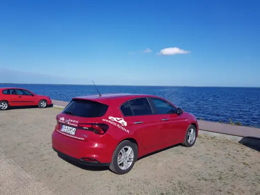 Czerwony fiat tipo sedan z napisem "SprawdzAuto24.pl" stoi nad błękitnym jeziorem w słoneczny dzień.