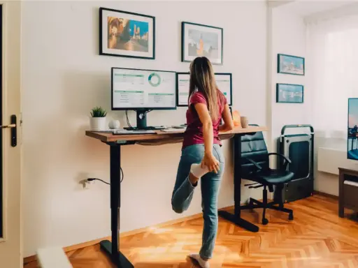 Femme s'étirant près de son bureau réglable en hauteur, pour un confort au bureau optimal. Deux écrans affichent des graphiques.