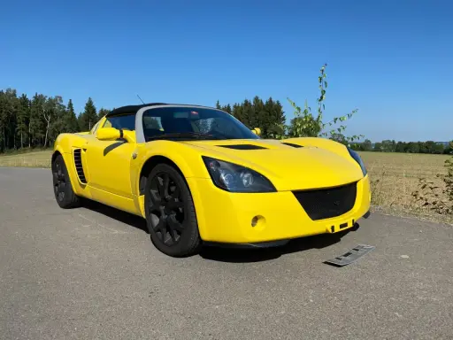 Żółty Opel Speedster na tle lasu i pól. Słońce pięknie podkreśla jego sportową sylwetkę.
