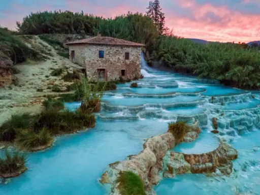 Cascate termali turchesi in Toscana, con un antico mulino in pietra e un cielo rosa al tramonto. Curiosità sulla Toscana: un luogo magico.