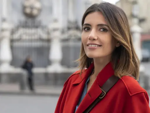 Donna con cappotto rosso sorride, con uno sfondo sfocato di edifici e una persona. Forse sta pensando ai **mina settembre libri** da leggere.