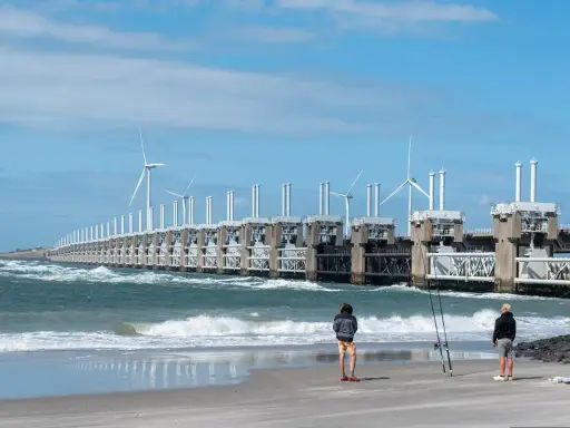 Dwie osoby stoją na plaży, obserwując falujące morze i imponującą zaporę z turbinami wiatrowymi. Takie morze jest w Holandii, chronione przez innowacyjne konstrukcje.