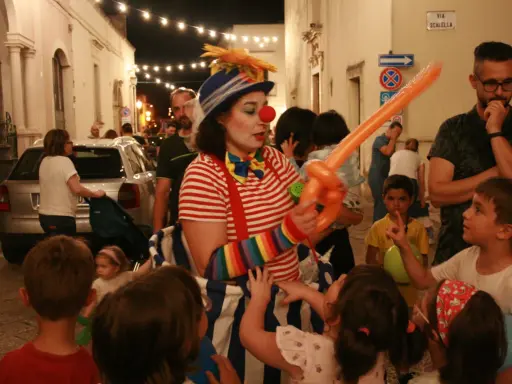 Clown con palloncino arancione anima la sagra di paese, circondata da bambini curiosi e adulti festanti.