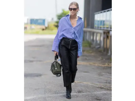 Donna con camicia a righe blu e bianche, occhiali da sole e un elegante outfit pantaloni ecopelle neri con zip frontale. Porta una borsa verde oliva.