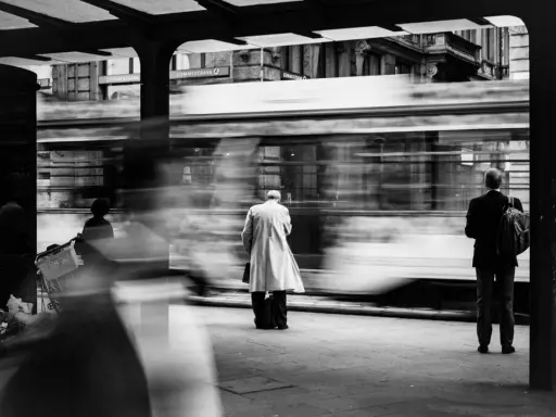 Street photography esempi: un tram sfreccia, lasciando figure sfocate in attesa alla fermata.