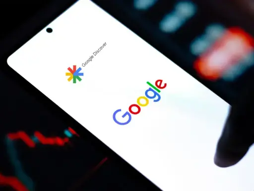 Ekran smartfona z logo Google i napisem Google Discover. W tle wykresy giełdowe.
