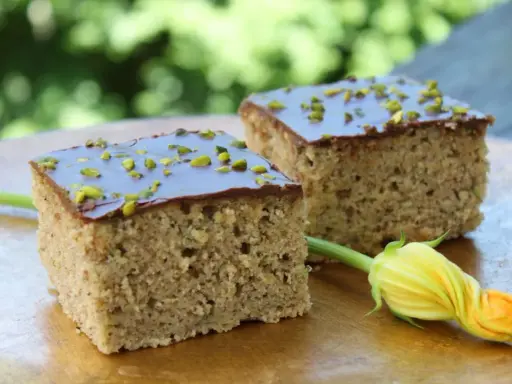 Zwei Stücke saftiger Zucchini Kuchen mit Schokoladenglasur und gehackten Pistazien, daneben eine Zucchiniblüte.