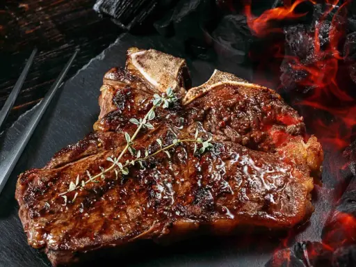 Un jugoso chuleton a la brasa, con romero fresco, listo para ser servido junto a brasas ardientes.