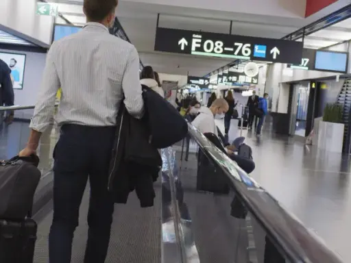 Resenärer på rullband på flygplatsen, skyltar visar vägen till gate F08-76. En man med resväskor på väg mot centrum.