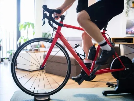 Intensywny trening na trenażerze kolarskim w domu. Czerwony rower, sportowiec w spodenkach i koszulce, butelki z wodą.