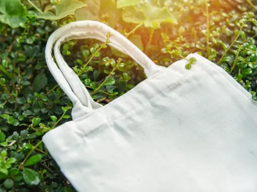Eine weiße Stofftasche liegt zwischen grünen Blättern. Perfekt, um die Einkaufsstaschen zu verstauen und die Umwelt zu schonen.
