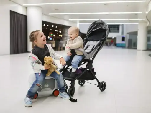 Uśmiechnięta dziewczynka z misiem siedzi na walizce obok wózka dla dziecka, w którym siedzi jej młodsze rodzeństwo.