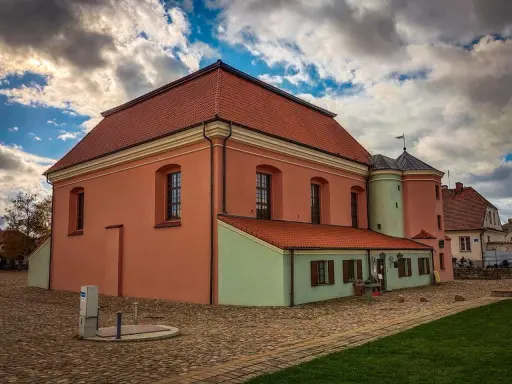 Tykocin co zobaczyć: zabytkowa Wielka Synagoga z różową fasadą i zielonym aneksem, na brukowanym placu pod burzliwym niebem.