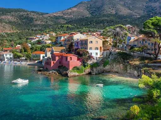 Case colorate affacciate su acque cristalline, un sogno di spiagge Grecia. Barche a vela e colline verdi completano il quadro.