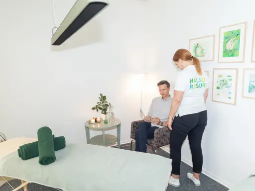 En kvinna i vit tröja med texten "HÄLSO RESURS" pratar med en man som sitter i en fåtölj. Rummet har en massagebänk och konst på väggarna, kanske för att förbereda för en vagusnerven massage.