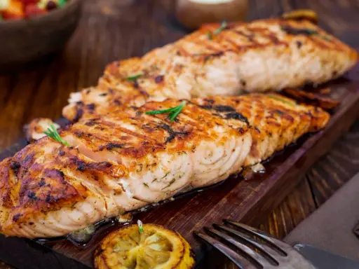Två saftiga laxfiléer, perfekt grillade på halster, ligger på en träbricka. En citronskiva och rosmarinkvistar pryder fisken.