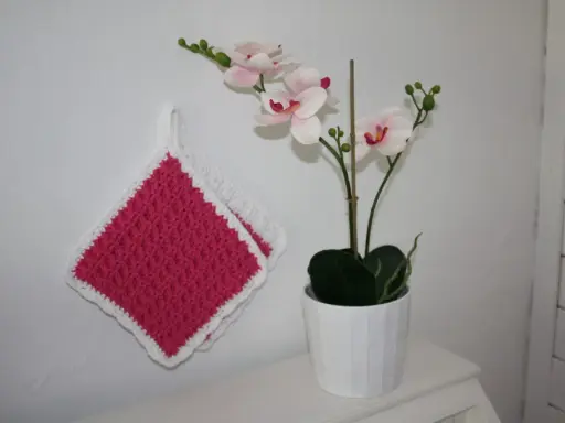 Två tjocka, virkade grytlappar i rosa med vit kant hänger på en vit vägg bredvid en orkidé i kruka.