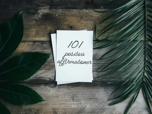 En hög med papper med texten "101 positiva affirmationer" på en träbakgrund, omgiven av gröna blad. En vacker påminnelse om affirmation svenska.