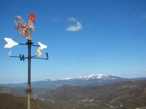 Gallo segnavento su sfondo azzurro, con montagne innevate all'orizzonte. Un invito a scoprire cosa vedere in Valmarecchia.