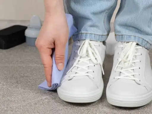Persona limpiando zapatillas blancas con un paño azul. A su lado, un cepillo y un bote de limpiador.
