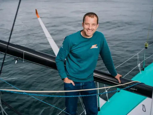 Sam Goodchild, souriant, sur un voilier, portant un sweat "For the Planet".