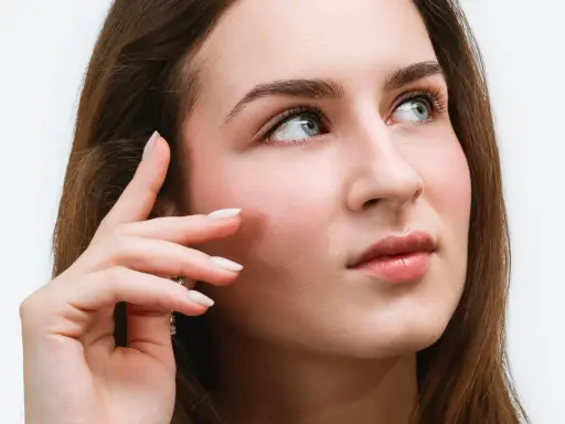 Une jeune femme aux yeux bleus regarde au loin, sa main effleurant sa joue. Ses cils, sublimés par un **tatouage cils**, accentuent son regard captivant.