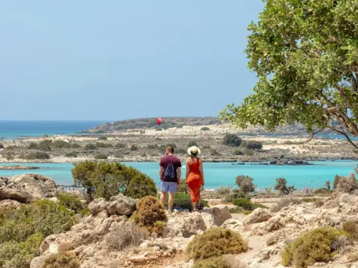 Ett par njuter av utsikten över Elafonissi, Kreta, med dess turkosa vatten och steniga landskap. En röd fallskärm syns i fjärran.