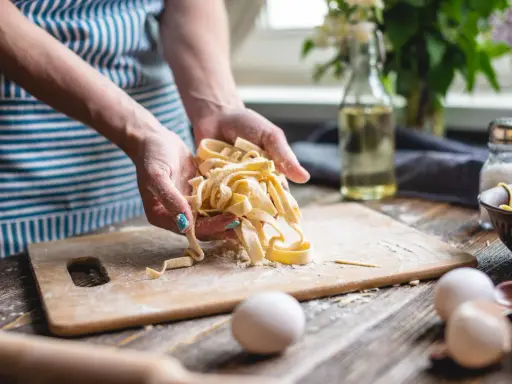 Hände halten frisch gemachte, italienische Pasta. Eier und Zutaten liegen bereit, um italienische Pasta selber machen zu können.