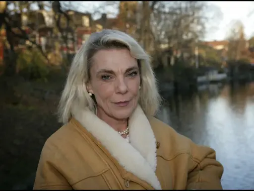 Pia Degermark idag, med sitt blonda hår och en varm fårskinnsjacka, står vid en vattensamling med träd och hus i bakgrunden.