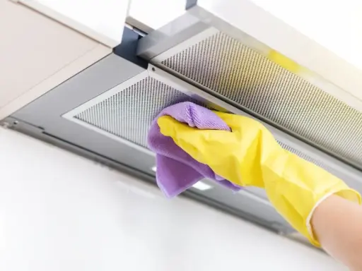 Mano con guante amarillo y paño morado limpiando la campana extractora por dentro.