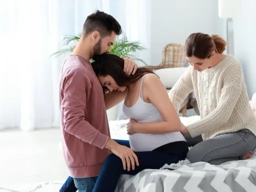Mężczyzna obejmuje ciężarną kobietę, która opiera głowę na jego ramieniu. Obok siedzi doula, która delikatnie dotyka jej brzucha, oferując wsparcie.